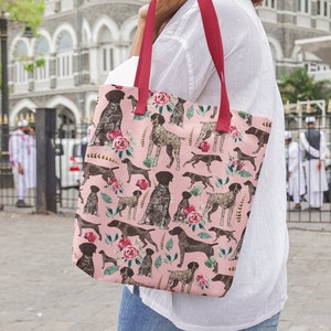 May include: A pink tote bag with a repeating pattern of brown and white German Shorthaired Pointers and pink roses. The bag has red handles.