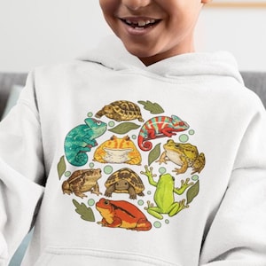 May include: White hooded sweatshirt featuring a colorful circular design of various reptiles and amphibians. The design includes a green chameleon, red and orange chameleon, frogs, and turtles. The sweatshirt is made of a soft material.