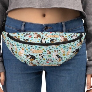 May include: A blue and white fanny pack with a pattern of Australian Shepherd dogs. The pack has a black adjustable strap and a black zipper.