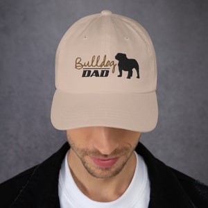 May include: A beige baseball cap with an embroidered black bulldog silhouette and the text "Bulldog DAD" in brown thread.