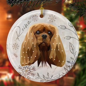 May include: A white ceramic Christmas ornament with a geometric illustration of a Cavalier King Charles Spaniel dog. The ornament has a white background with a gray marble pattern and a gray floral design. The dog's name, "Reina", is written in gray script on the ornament.