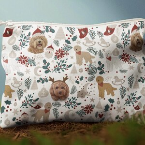 May include: A white zippered pouch with a repeating pattern of brown dogs, Christmas trees, Santa hats, and holly. The design includes red, green, and gray accents, creating a festive holiday theme. The pouch is sitting on a bed of green moss.