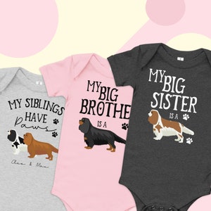 Op de afbeelding: Drie baby-rompers met verschillende hondenthema-ontwerpen. De eerste romper is grijs met de tekst "Mijn broertjes en zusjes hebben pootjes" en de namen "Cleo & Max". De tweede romper is roze met de tekst "Mijn grote broer is een" en een zwarte Cavalier King Charles Spaniel. De derde romper is grijs met de tekst "Mijn grote zus is een" en een bruine Cavalier King Charles Spaniel.