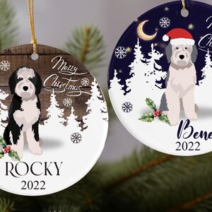 Op de afbeelding: Twee ronde kerstversieringen met hondenillustraties. Eén heeft een zwart-witte hond op een houten achtergrond met de naam "Rocky" en het jaar "2022". De andere heeft een hond met een kerstmuts, de naam "Benett" en het jaar "2022". Beide zeggen "Merry Christmas".
