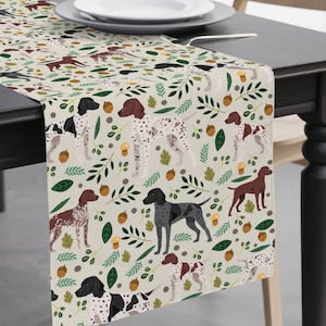 May include: A beige table runner with a repeating pattern of brown, black, and white hunting dogs, green leaves, and brown acorns.