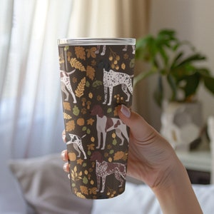 May include: A brown and white insulated tumbler with a pattern of German Shorthaired Pointers and autumn leaves. The tumbler has a clear lid.
