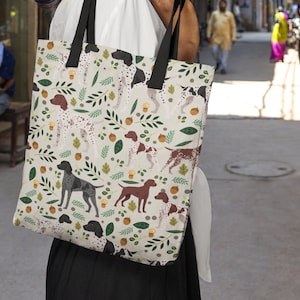 Puede incluir: Una bolsa de tela blanca con asa negra y un patrón de perros marrones y blancos, hojas verdes y bellotas marrones.