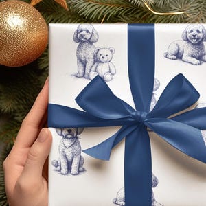 Puede incluir: Un regalo envuelto con papel de regalo blanco con bocetos azules de caniches y un oso de peluche. El regalo está atado con una cinta y un lazo de satén azul marino. Una bola dorada y ramas de coníferas están en el fondo.