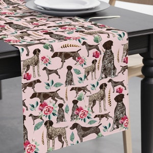 May include: A pink table runner featuring a repeating pattern of German Shorthaired Pointers, roses, and greenery. The dogs are depicted in various poses, with brown and white speckled coats. The runner is draped over a dark table, with a white plate and silverware visible.