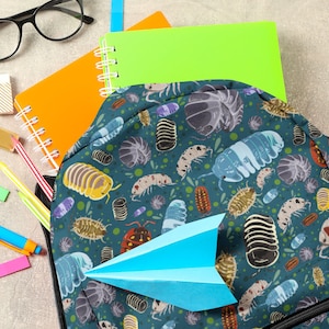 May include: A blue backpack with a colorful pattern of isopods, or pill bugs, on a green background. The backpack is partially unzipped and a blue paper airplane is sticking out of the opening.