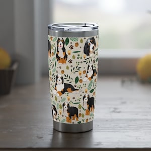 May include: A stainless steel travel mug with a white background and a pattern of Bernese Mountain Dogs, leaves, acorns, and paw prints.