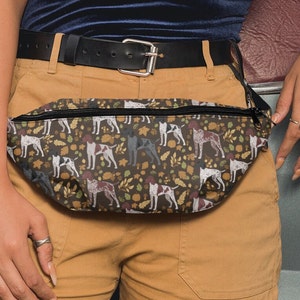 May include: A brown and black fanny pack with a zipper closure. The fanny pack has a pattern of brown and white dogs with autumn leaves and acorns.