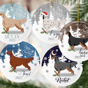 May include: Set of five round ceramic Christmas ornaments featuring a winter scene with a dog breed, each with a different dog name and the year 2022. The ornaments are white with a blue, gray, or brown background. The dogs are a white and brown English Setter, a black and tan Irish Setter, a black and white English Setter, a black and tan Gordon Setter, and a black and white English Setter.