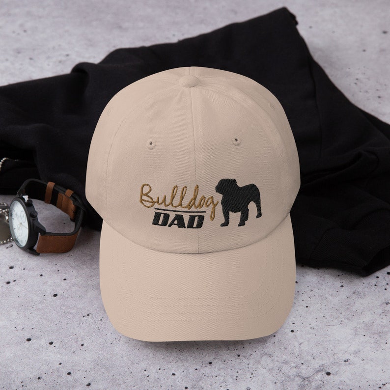 May include: A beige baseball cap with an embroidered black bulldog silhouette and the text "Bulldog DAD" in brown thread.