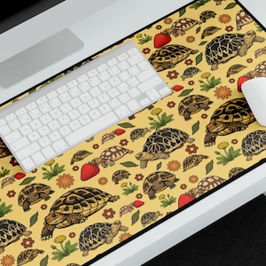 Puede incluir: Alfombrilla de escritorio amarilla con un patrón repetido de tortugas, fresas y flores. Un teclado y un ratón blancos están sobre la alfombrilla. La alfombrilla es rectangular con un borde negro.