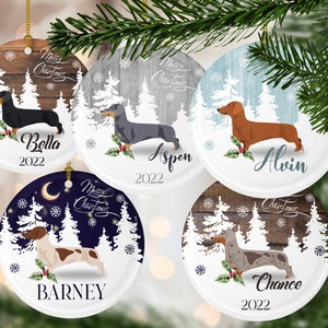 May include: Five round, white, ceramic Christmas ornaments with a winter woodland scene and a dachshund dog breed. Each ornament features a different colored background and a different dachshund dog breed. The ornaments are personalized with the names Bella, Aspen, Alvin, Barney, and Chance, and the year 2022.