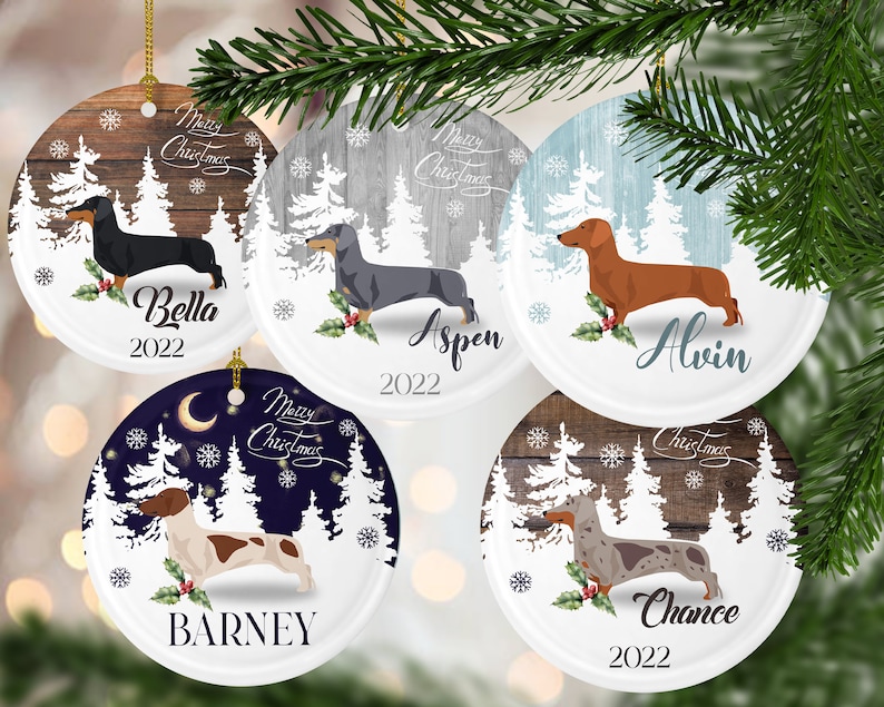May include: Five round, white, ceramic Christmas ornaments with a winter woodland scene and a dachshund dog breed. Each ornament features a different colored background and a different dachshund dog breed. The ornaments are personalized with the names Bella, Aspen, Alvin, Barney, and Chance, and the year 2022.