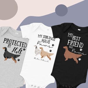 May include: Three baby onesies with dog illustrations and text. The grey onesie says "Protected by Holt" with a black dog illustration. The white onesie says "My siblings have paws" with two dog illustrations and the names "Blits & Basil". The black onesie says "My best friend has paws" with a brown dog illustration.