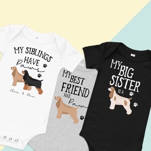 Puede incluir: Tres bodies de bebé con diferentes diseños de temática canina. El body blanco dice "Mis hermanos tienen patas" con dos perros, uno negro y uno marrón. El body gris dice "Mi mejor amigo tiene patas" con un perro marrón. El body negro dice "Mi hermana mayor es una pata" con un perro marrón y blanco.