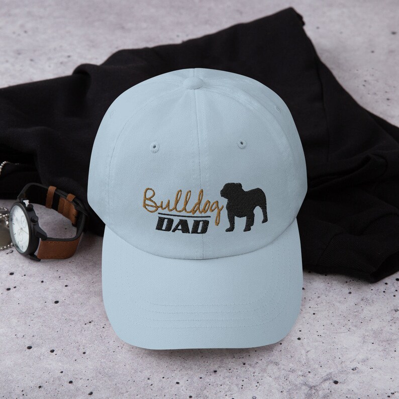 May include: A light blue baseball cap with a black bulldog silhouette and the text "Bulldog Dad" embroidered in gold thread.