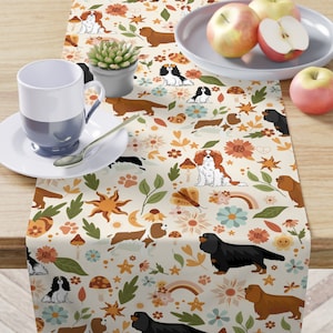 May include: A beige table runner with a repeating pattern of Cavalier King Charles Spaniels, flowers, butterflies, rainbows, and stars. The dogs are brown, black, and white. The flowers are pink, yellow, orange, and blue. The background is a light beige.