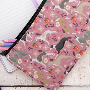 May include: A pink zippered pencil case with a repeating pattern of greyhounds, strawberries, and flowers. The case is partially open and has several colored pencils inside.