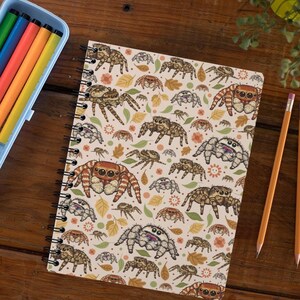 May include: A spiral-bound notebook featuring a repeating pattern of various jumping spiders in shades of brown, orange, and white, interspersed with small autumn-colored leaves and orange flower icons on a light cream-colored background.