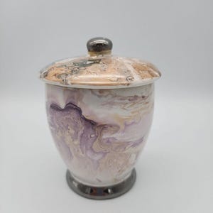 May include: A decorative ceramic jar with a lid. The jar features a swirling pattern of purple, white, and beige. The lid has a metallic silver knob and a similar marbled design. The base of the jar is also silver.