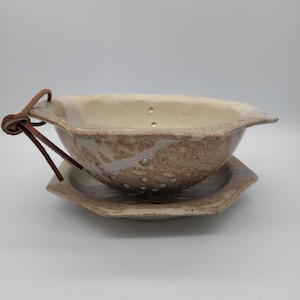 May include: A handmade ceramic colander with a matching saucer. The colander is a speckled brown and beige with drainage holes. It has a leather tie attached to the rim. The saucer is a matching speckled beige.