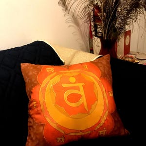 May include: Orange and brown decorative pillow with a large, stylized yellow symbol at the center. The pillow has a floral design around the symbol and is placed on a black surface. The pillow is likely part of a home decor collection.