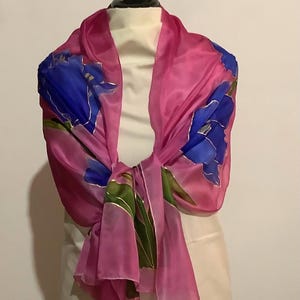 May include: A pink silk scarf with a floral design. The scarf features large blue flowers with gold accents and green leaves. The scarf is draped over a mannequin, showcasing its flowing fabric and vibrant colors.