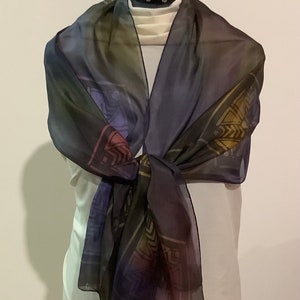 Op de afbeelding: Een transparante zijden sjaal met een geometrisch patroon in tinten van paars, groen en goud. De sjaal is om de nek van een mannequin geknoopt.