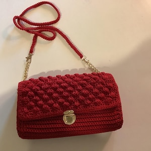 May include: A red crochet crossbody bag with a textured, raised pattern. The bag features a flap closure with a gold-colored clasp and a matching gold chain strap. The strap is attached to the bag with gold-colored hardware.