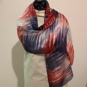 Pode incluir: Uma longa echarpe fluida com um padrão abstrato em vermelho, branco e azul. A echarpe é feita de um tecido macio e transparente e está drapeada sobre um manequim branco.
