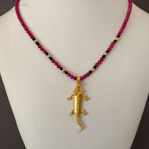 Könnte beinhalten: Eine Halskette mit einem goldenen Eidechsen-Anhänger und einer Kette aus rosa und schwarzen Perlen.