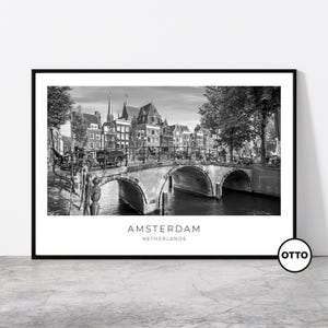 May include: Monochrome framed print depicting a canal bridge in Amsterdam, Netherlands. The image shows a stone bridge with arched openings, bicycles, and buildings. The word "AMSTERDAM" is printed at the bottom of the print.