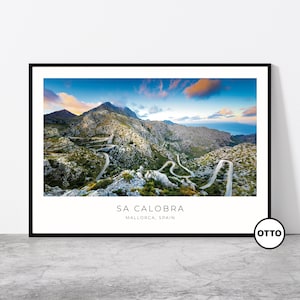 May include: A framed print depicting the Sa Calobra road in Mallorca, Spain. The photograph captures a winding road through a mountainous landscape, set against a backdrop of a blue sky and clouds. The text "SA CALOBRA MALLORCA, SPAIN" is visible.