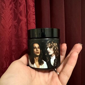 May include: A black glass jar with a label featuring two men with long hair. The men are wearing white shirts and have a dark background.