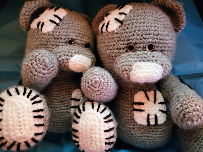 May include: Two gray crocheted teddy bears with white patches and black accents. The bears have black button eyes and white noses.