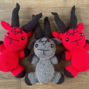 May include: Three crocheted plush toys shaped like goats with horns. Two are red with black horns and a pentagram on their chests. One is grey with black horns and a pentagram on its chest.