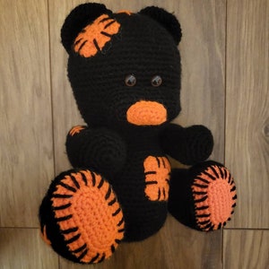 May include: A black crocheted teddy bear with orange accents. The bear has a stitched-on orange nose and brown eyes. The bear's paws and ears are accented with orange crocheted details.