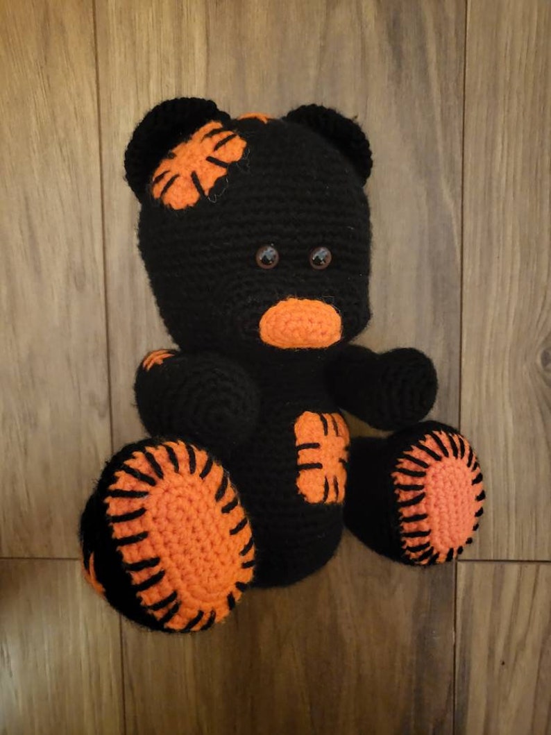 May include: A black crocheted teddy bear with orange accents. The bear has a stitched-on orange nose and brown eyes. The bear's paws and ears are accented with orange crocheted details.