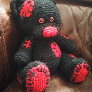 May include: A black crocheted teddy bear with red accents. The bear has red eyes and a red nose. The bear is sitting on a brown leather couch.