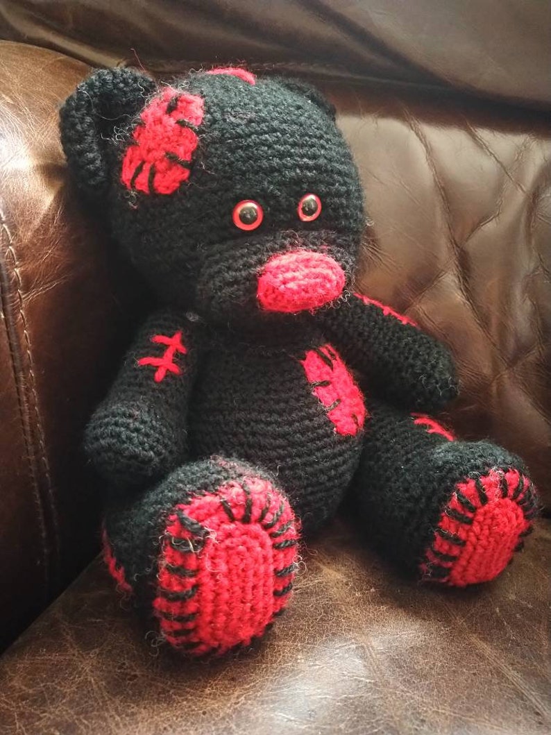 May include: A black crocheted teddy bear with red accents. The bear has red eyes and a red nose. The bear is sitting on a brown leather couch.