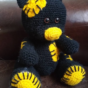 May include: A black crocheted teddy bear with yellow accents. The bear has a stitched-on yellow nose and yellow patches on its head, arms, and feet.