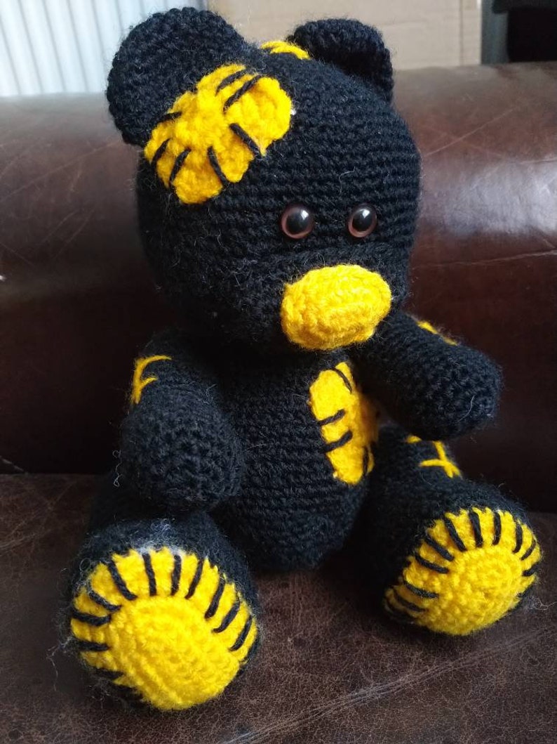 May include: A black crocheted teddy bear with yellow accents. The bear has a stitched-on yellow nose and yellow patches on its head, arms, and feet.