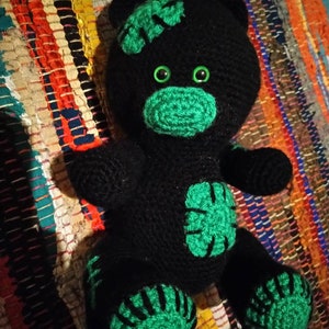 May include: Black crocheted teddy bear with green accents. The bear has large green eyes and a green nose. The bear's paws and ears are also green.