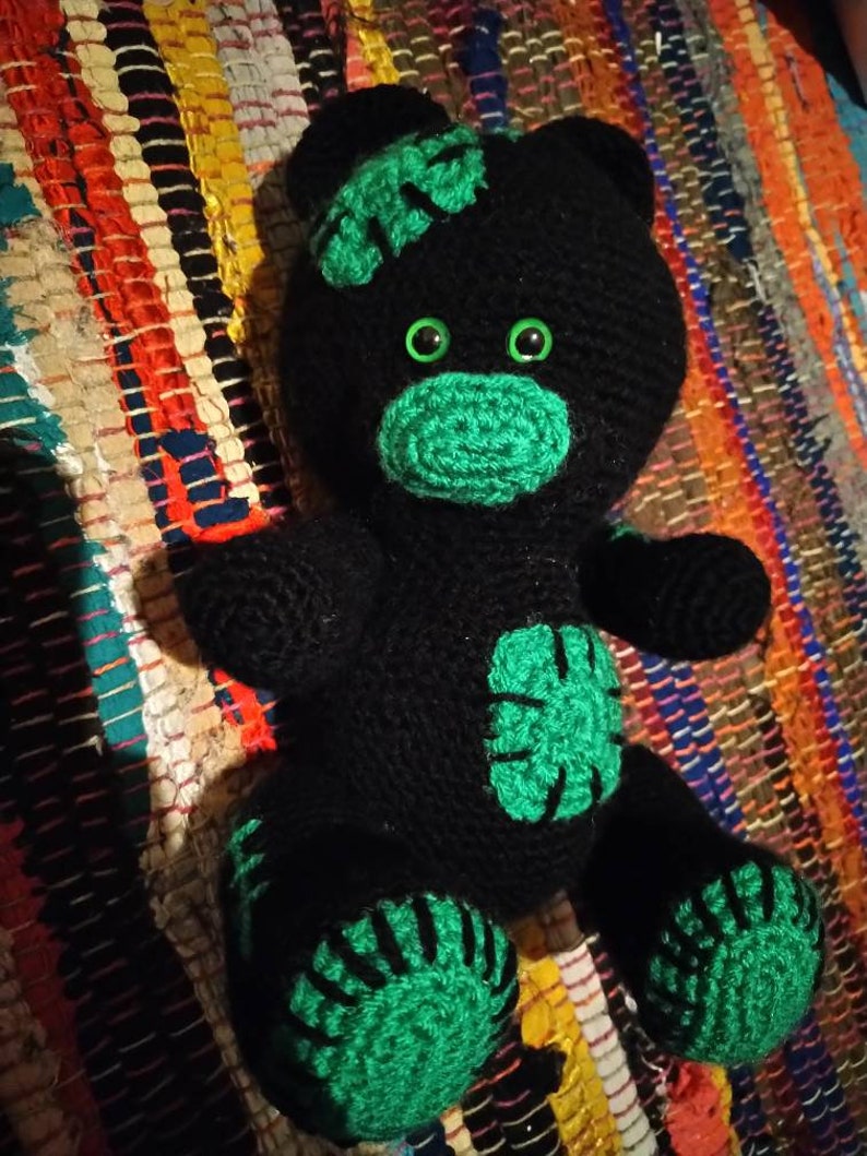 May include: Black crocheted teddy bear with green accents. The bear has large green eyes and a green nose. The bear's paws and ears are also green.