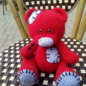 May include: A red crocheted teddy bear with gray patches and black stitching. The bear is sitting on a brown and white wicker chair.