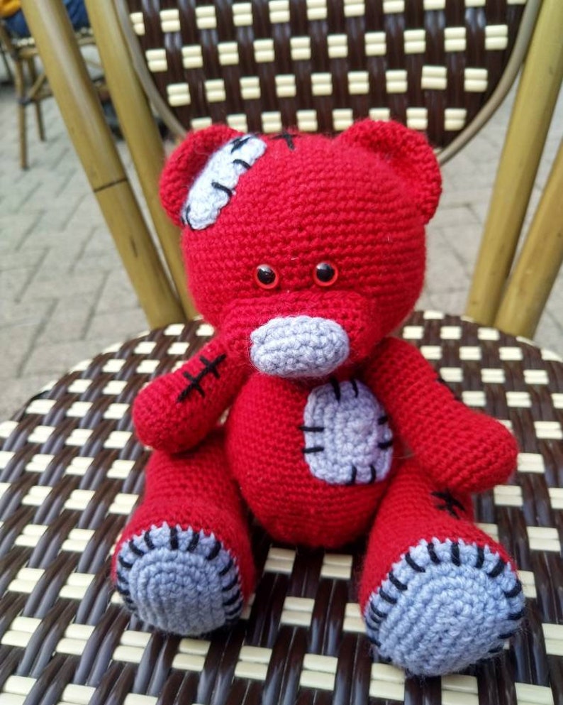 May include: A red crocheted teddy bear with gray patches and black stitching. The bear is sitting on a brown and white wicker chair.